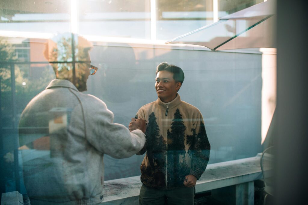 Two men smile and shake hands through a glass window, reflecting a warm friendly interaction.