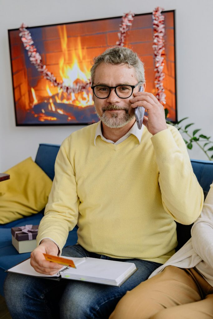 pexels-photo-5810697-5810697 Elderly man with glasses sitting by the fireplace, talking on the phone and smiling warmly.