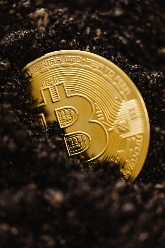 pexels-photo-5980916-5980916-1 Close-up of a gold Bitcoin coin emerging from dark soil, representing digital currency growth.