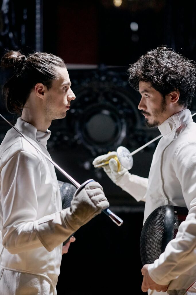 Two fencers in traditional gear engage in a tense face-off in an indoor setting.