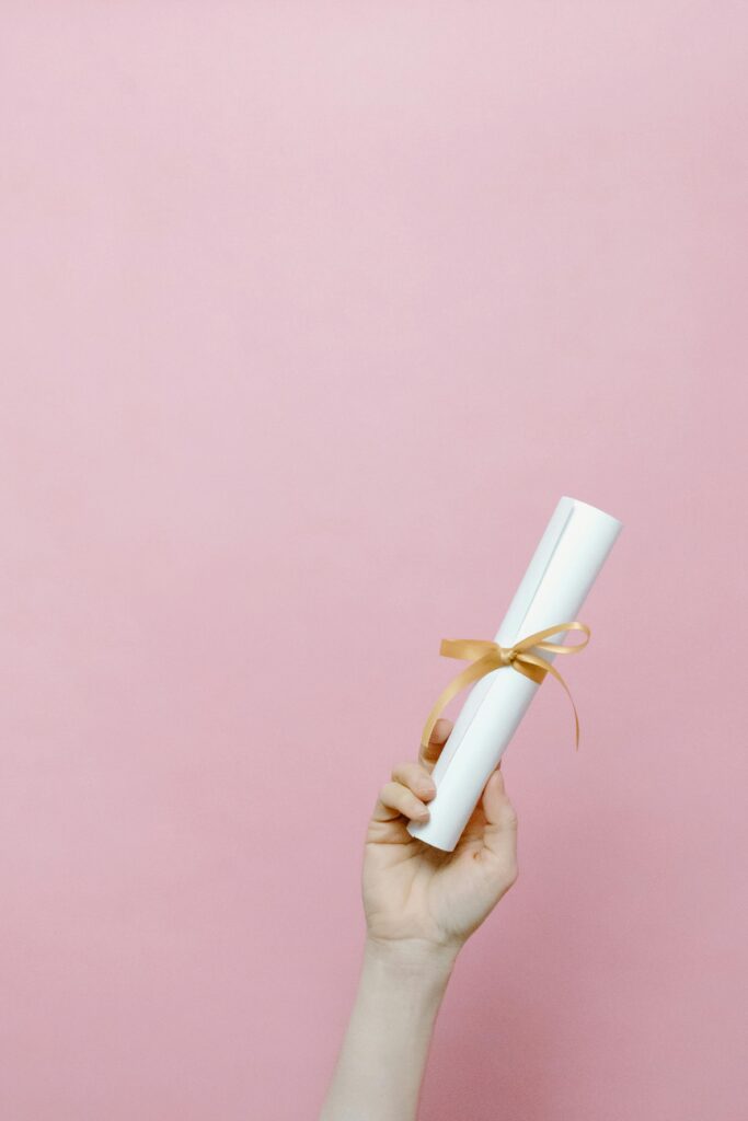 pexels-photo-7723829-7723829 A hand holding a rolled diploma tied with a ribbon against a pink background.